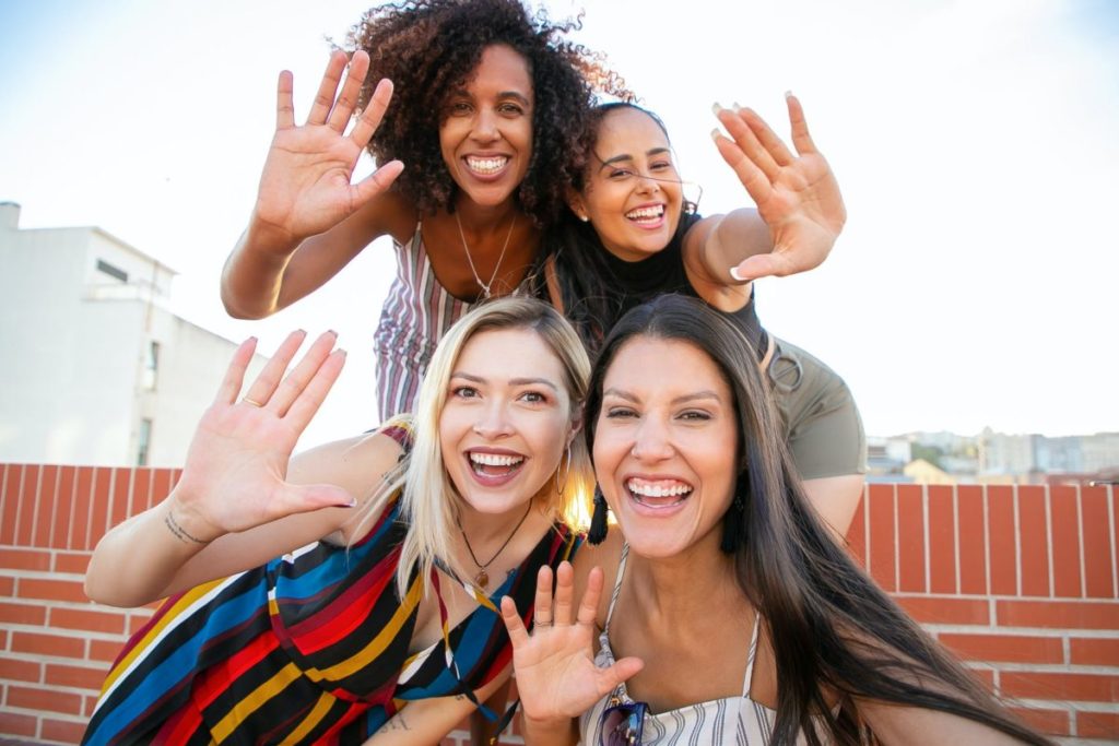 smiling young diverse ladies showing hi sign while taking selfie on terrace